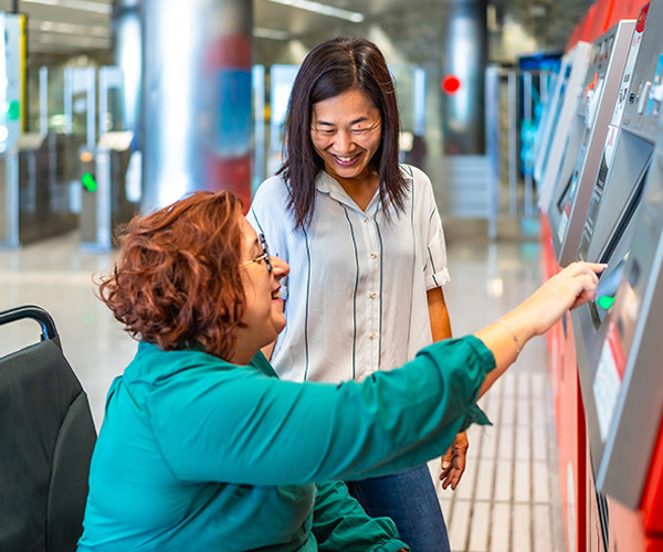 Woman in a wheelchair using a self-service terminal with the help of a smiling assistant, in a public environment