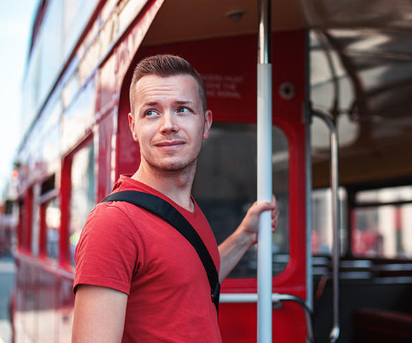 Young man in red t-shirt and black backpack gets on an iconic red double-decker bus, while looking to the side with a serene expression